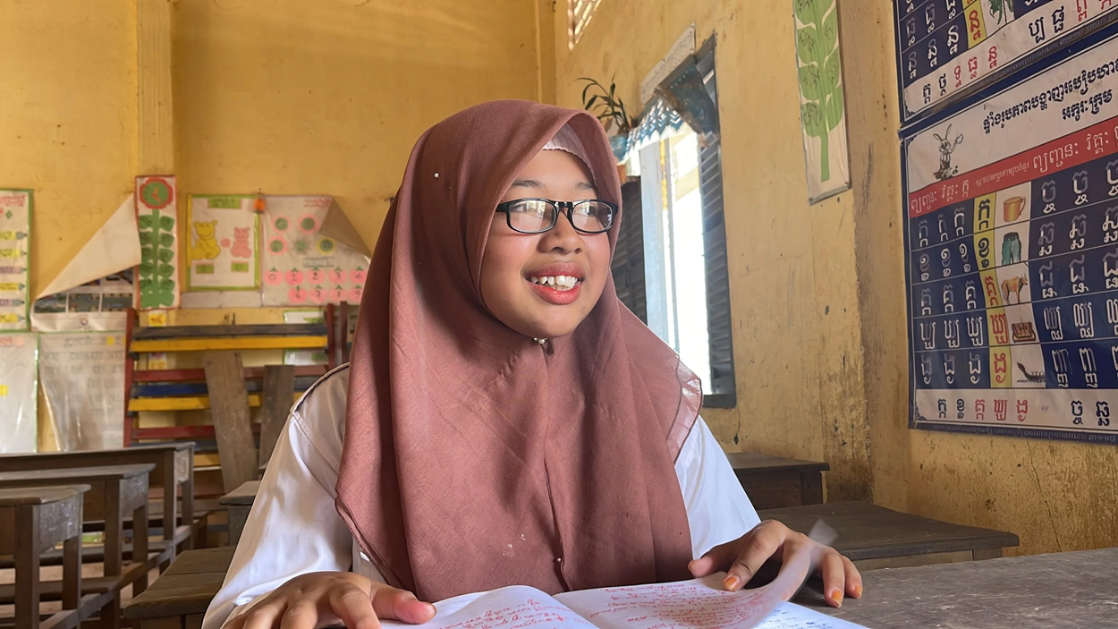 Schoolgirl wearing glasses and a headscarf sits at a classroom desk, smiling while reading from an open book