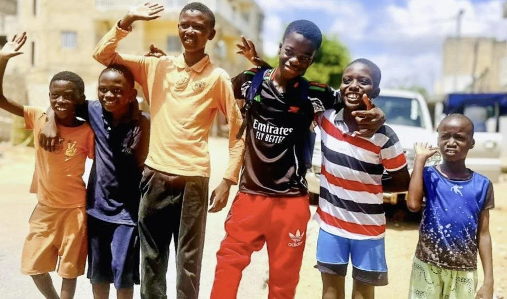 Six children stand together outdoors, smiling, waving, and posing with their arms around each other in a neighbourhood street.