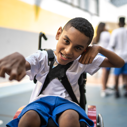 A smiling school-aged child using a wheelchair gestures playfully in a school setting