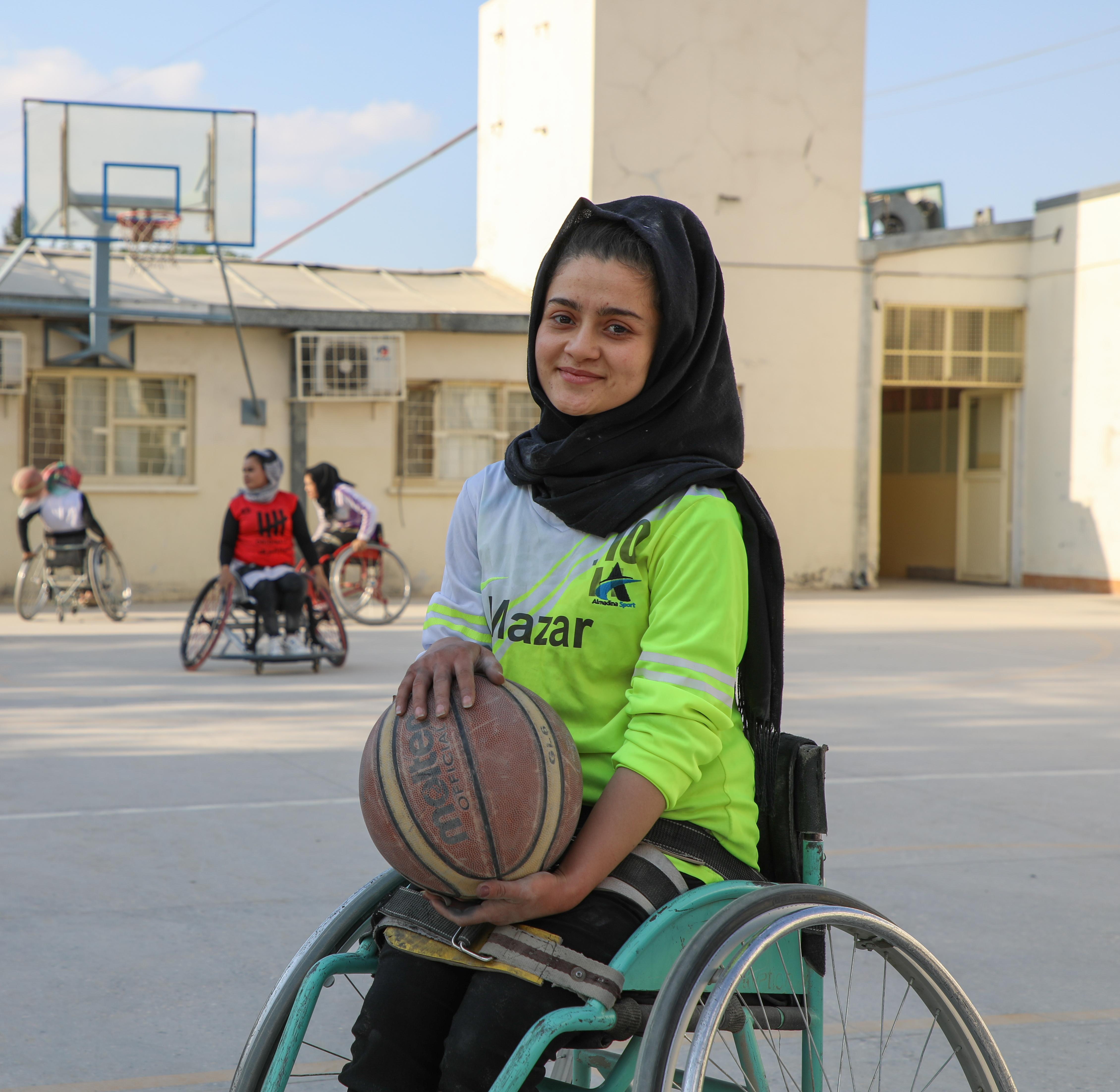A young woman using a wheelchair holds a basketball on an outdoor court, with others playing wheelchair basketball in the background