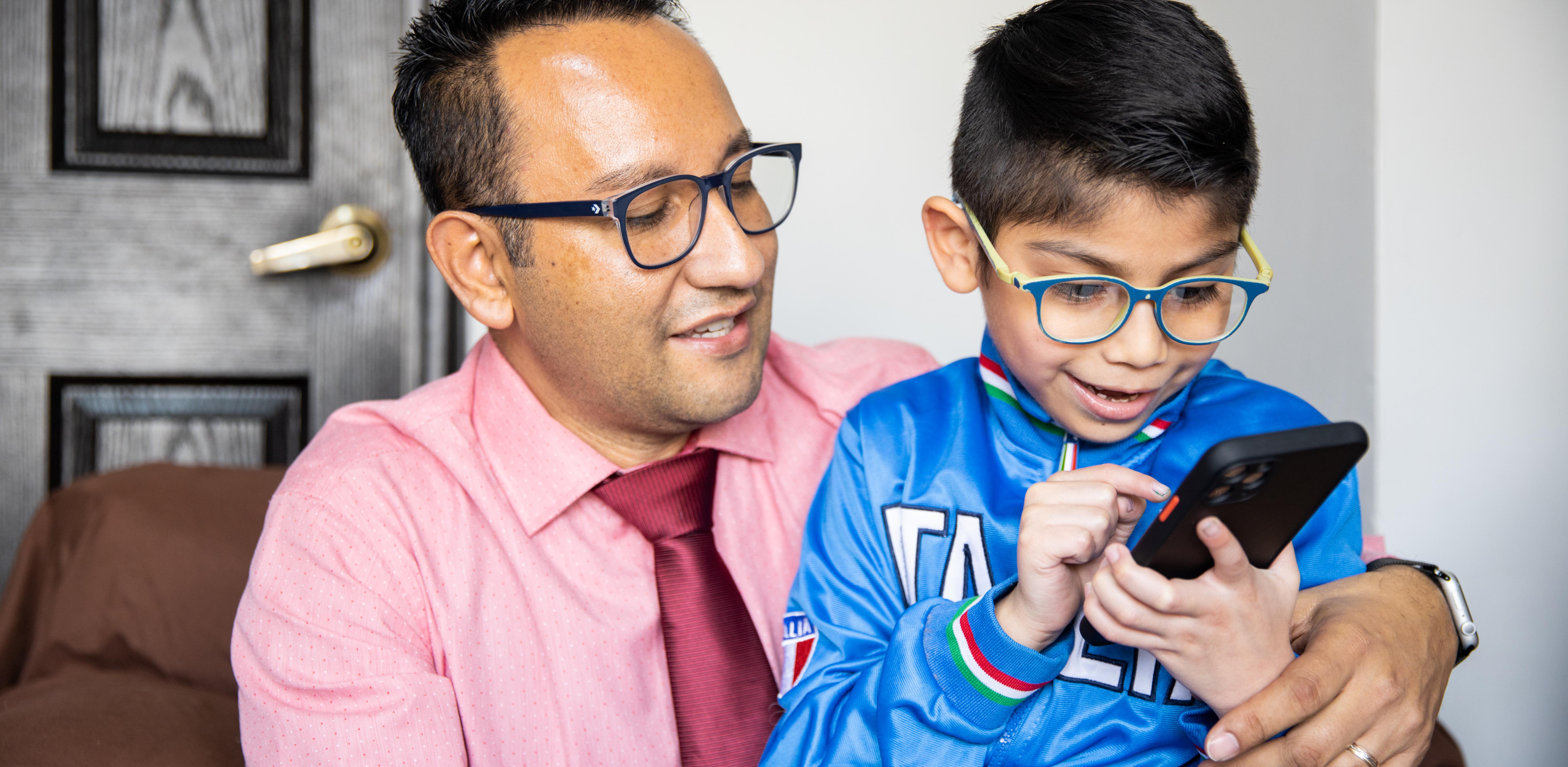 An adult sits with a child who is using a smartphone, both wearing glasses, indoors