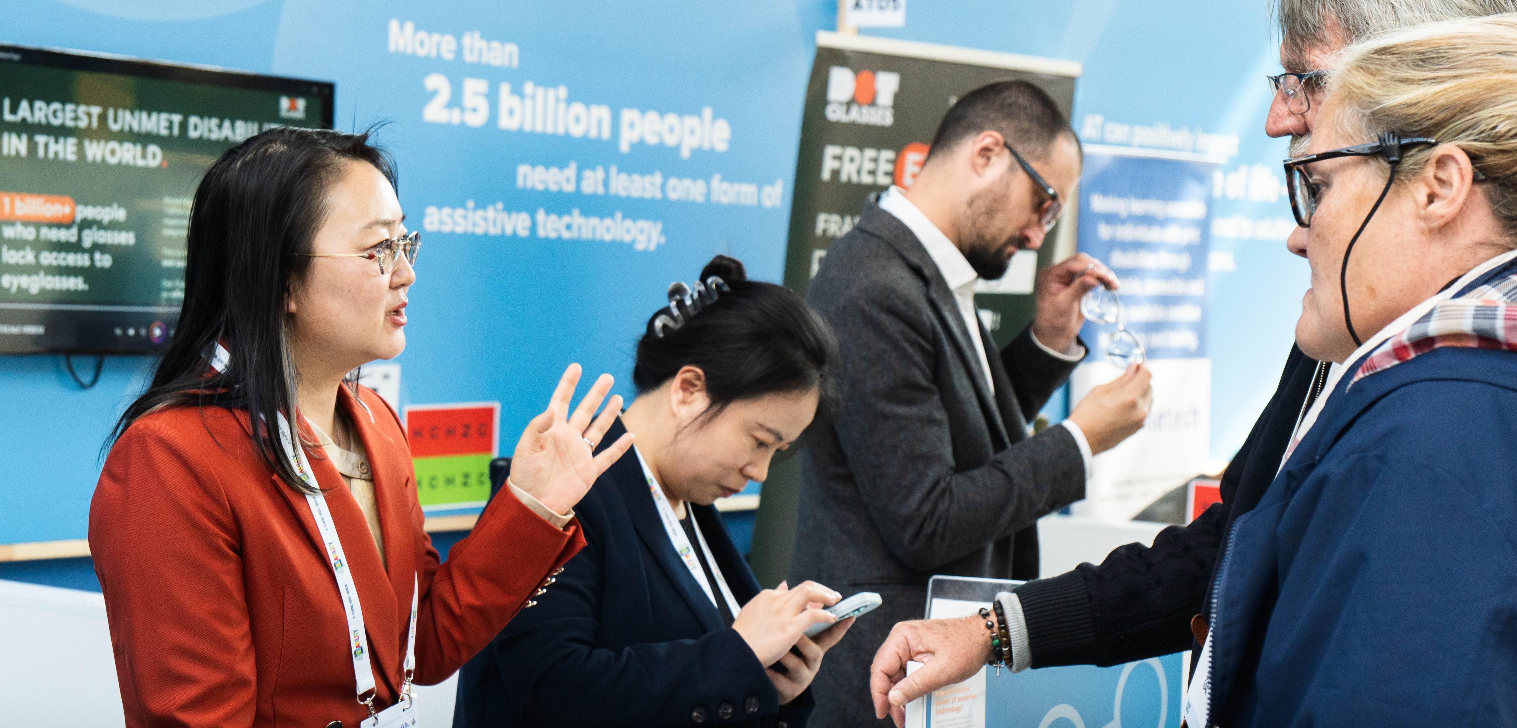 People discuss assistive technology products at an exhibition booth, with informational displays in the background.