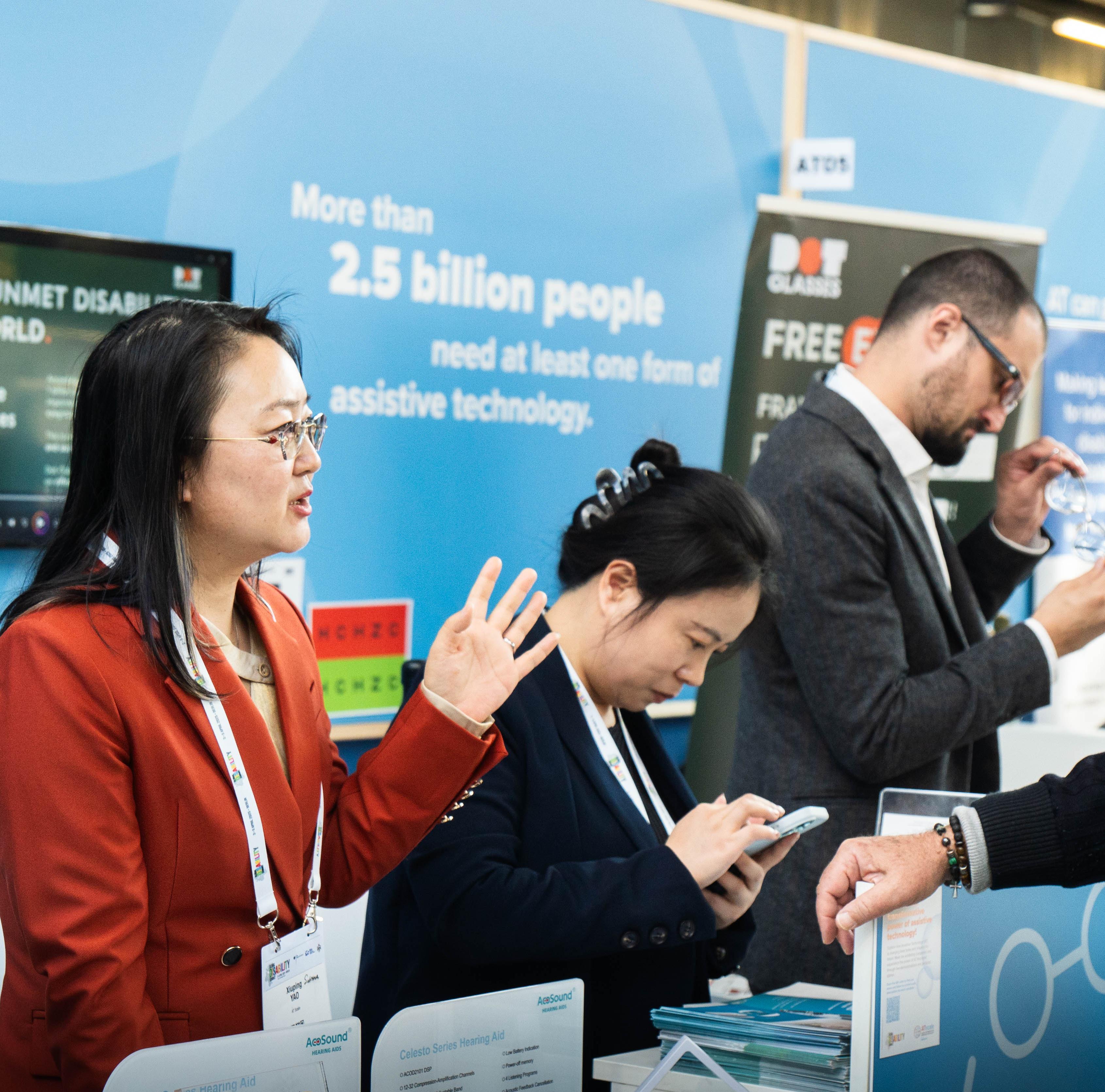 People discuss assistive technology products at an exhibition booth, with informational displays in the background
