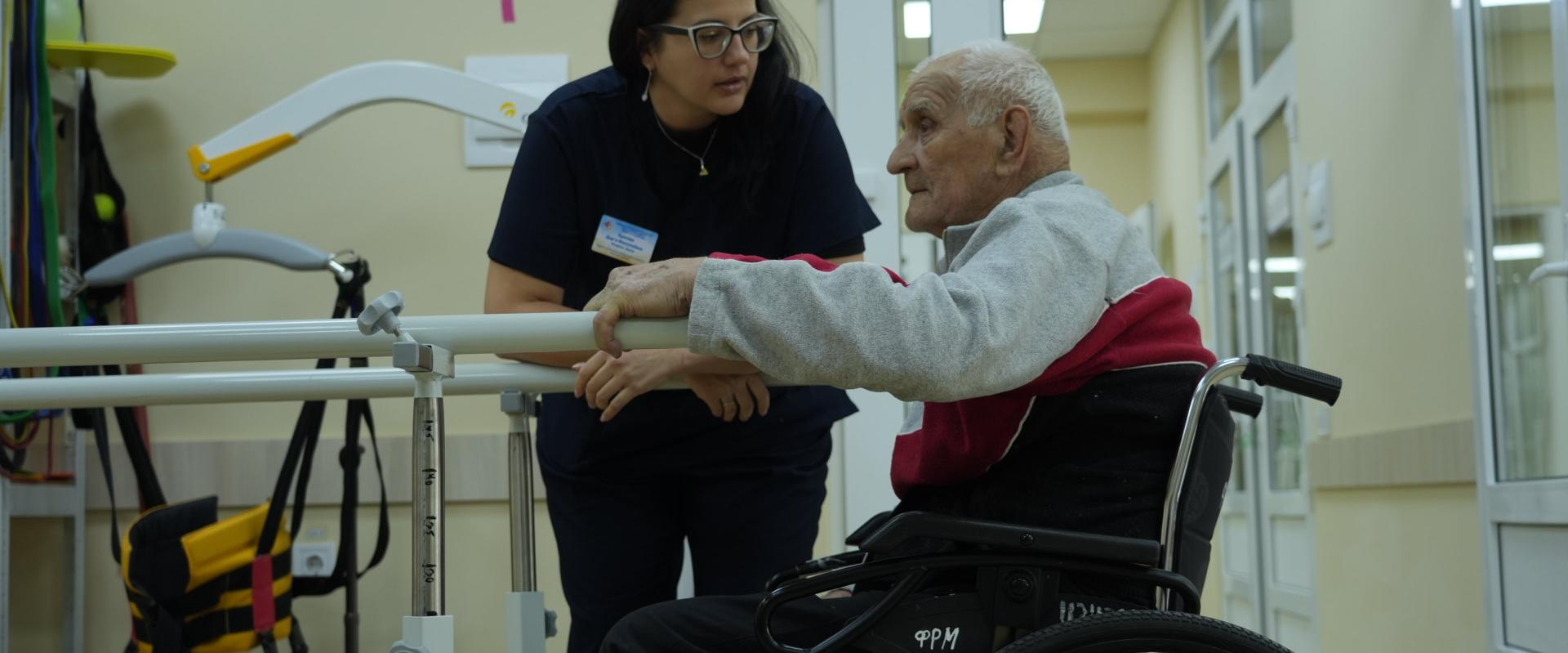 A health professional supports an older man using a wheelchair as he practices standing at parallel bars in a clinic.