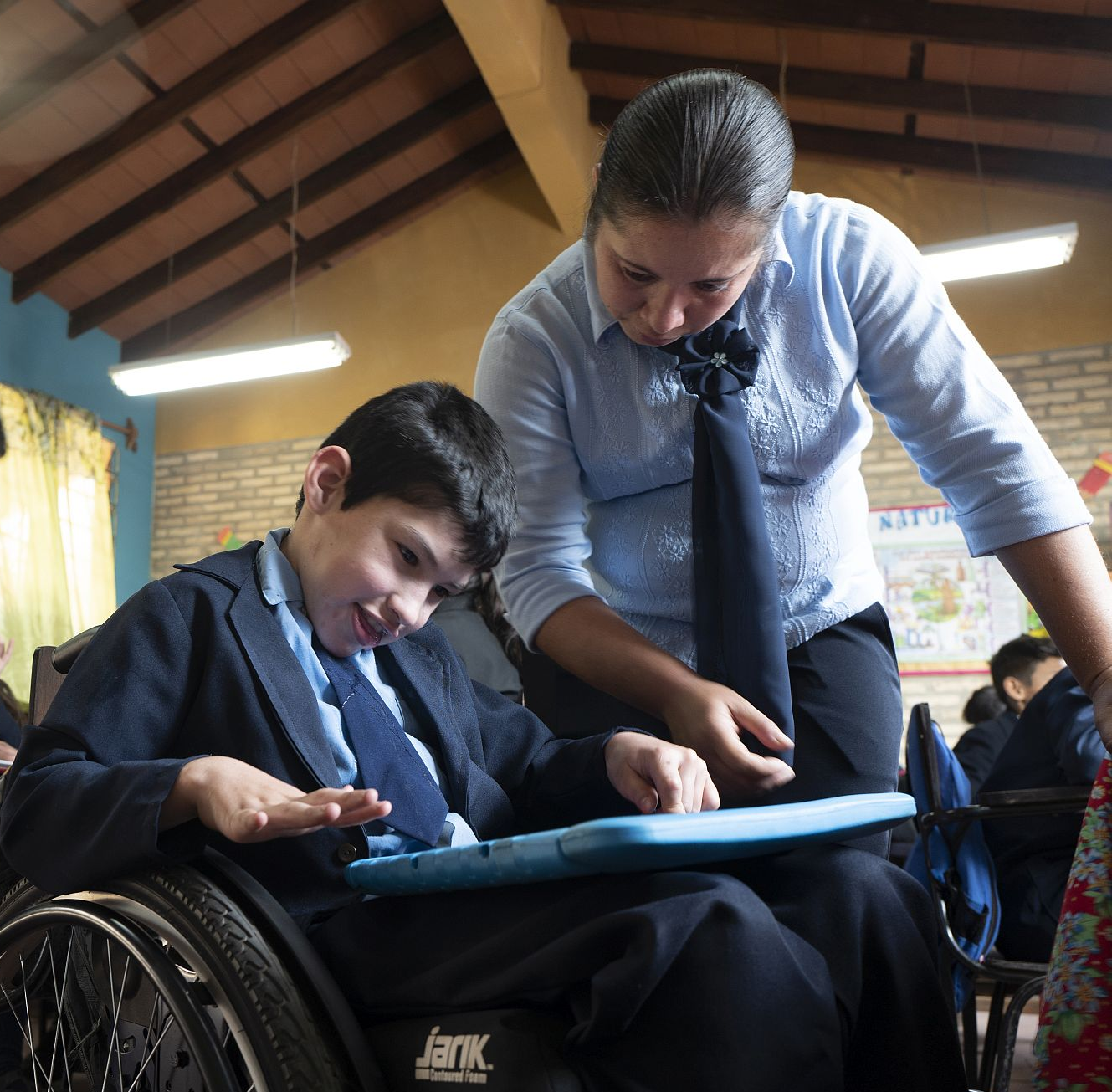 A teacher assists a young student using a tablet in a classroom.