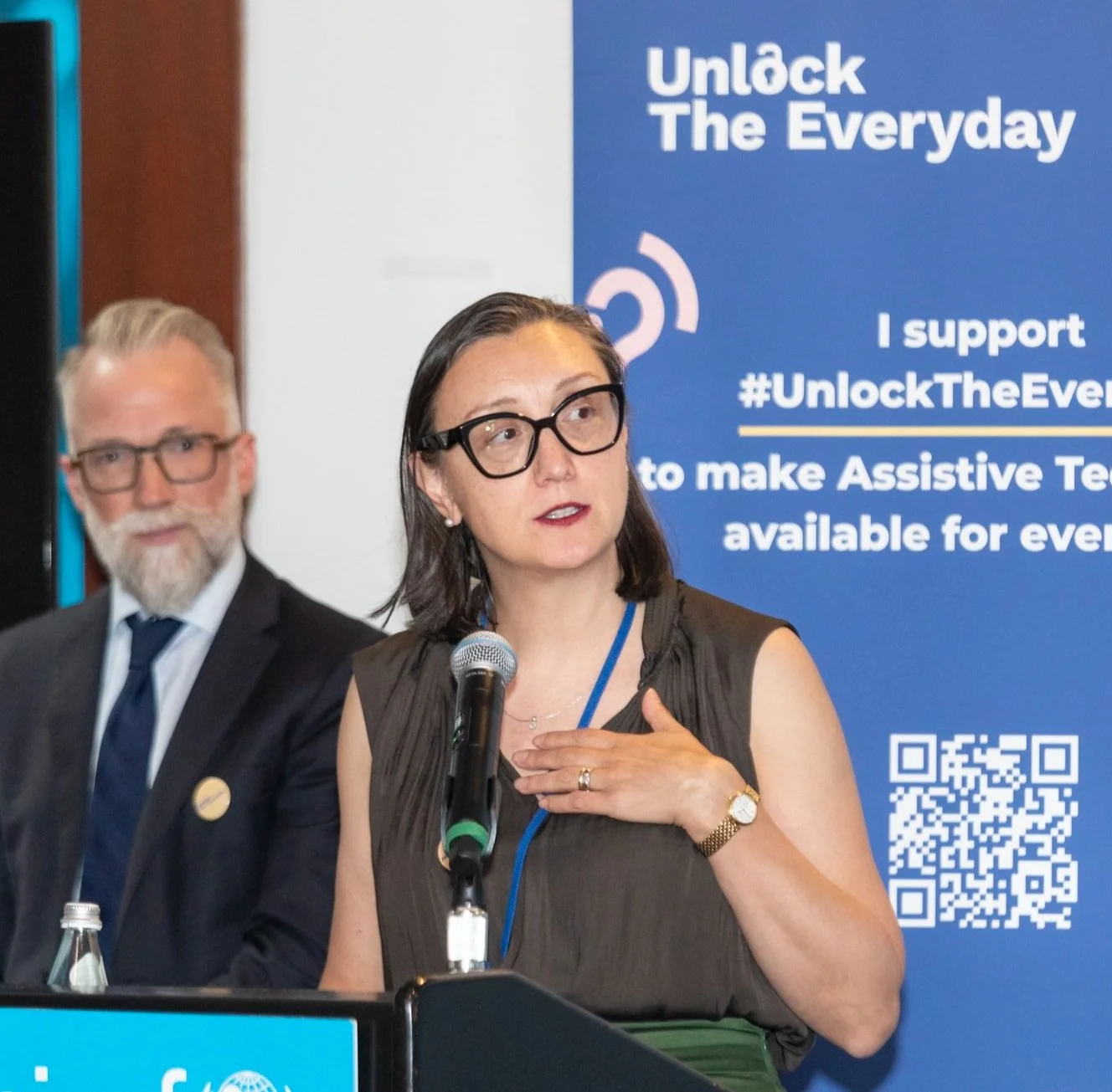 A woman speaks at a podium during an event, with a banner about assistive technology behind her and another speaker seated nearby