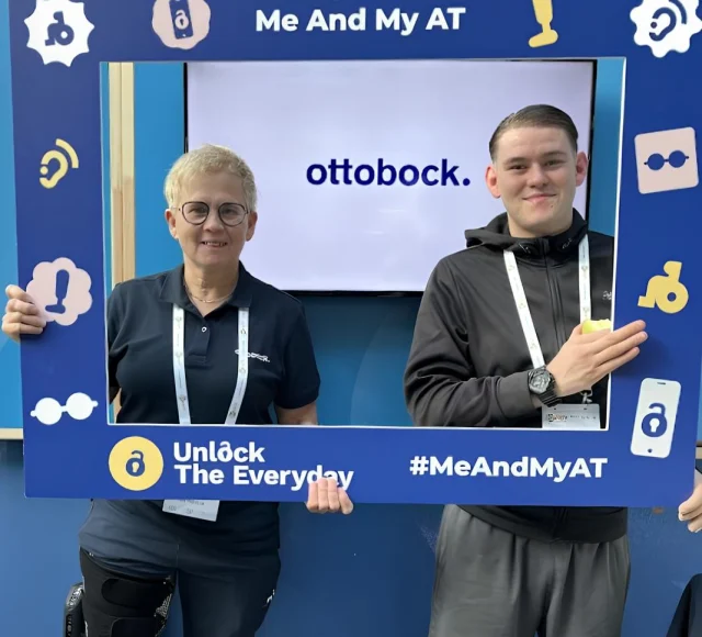 Two people pose indoors holding a ‘Me and My AT’ campaign frame, with ‘Unlock the Everyday’ and ‘#MeAndMyAT’ text and the Ottobock logo in the background.