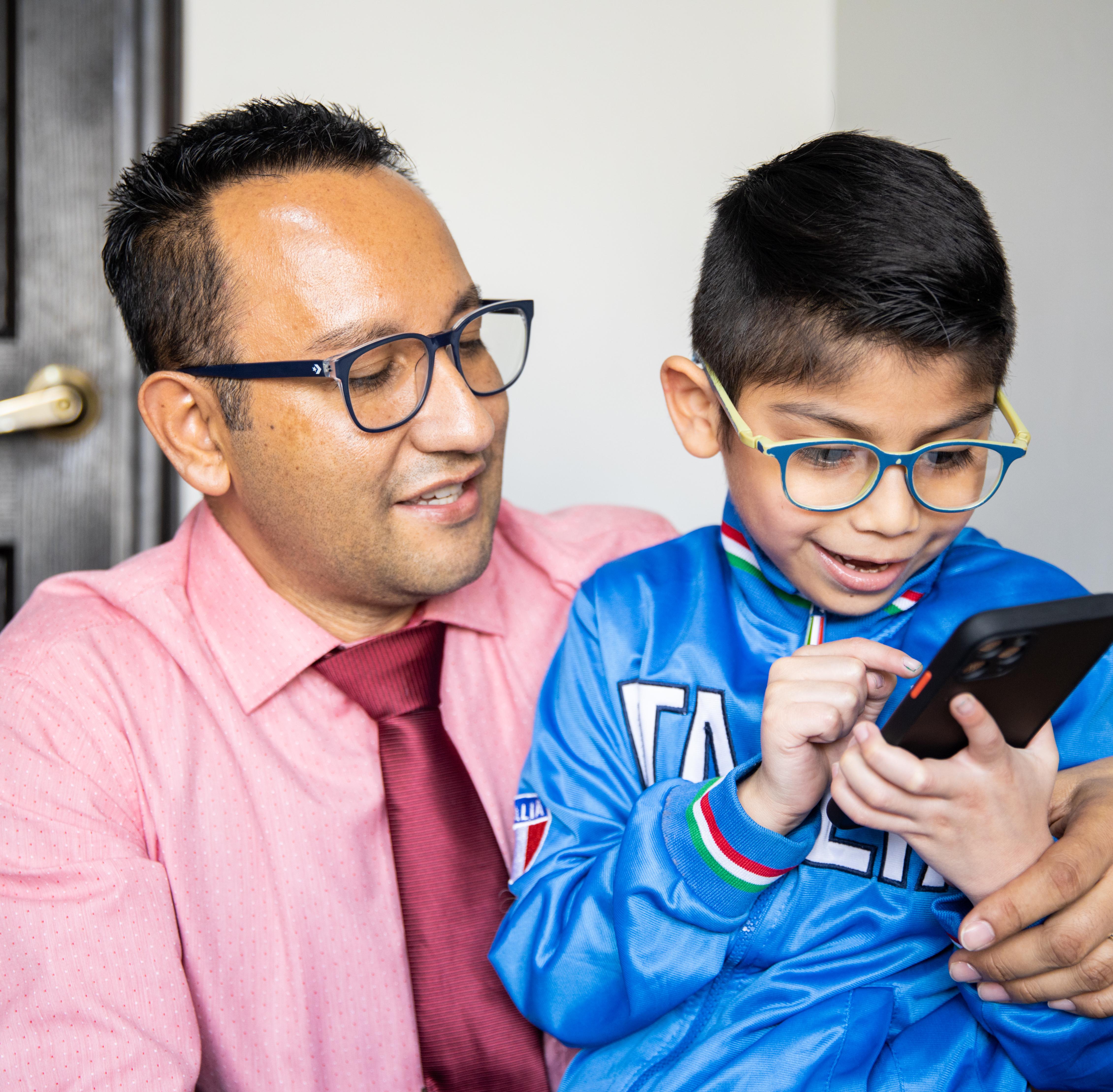 An adult sits with a child who is using a smartphone, both wearing glasses, indoors