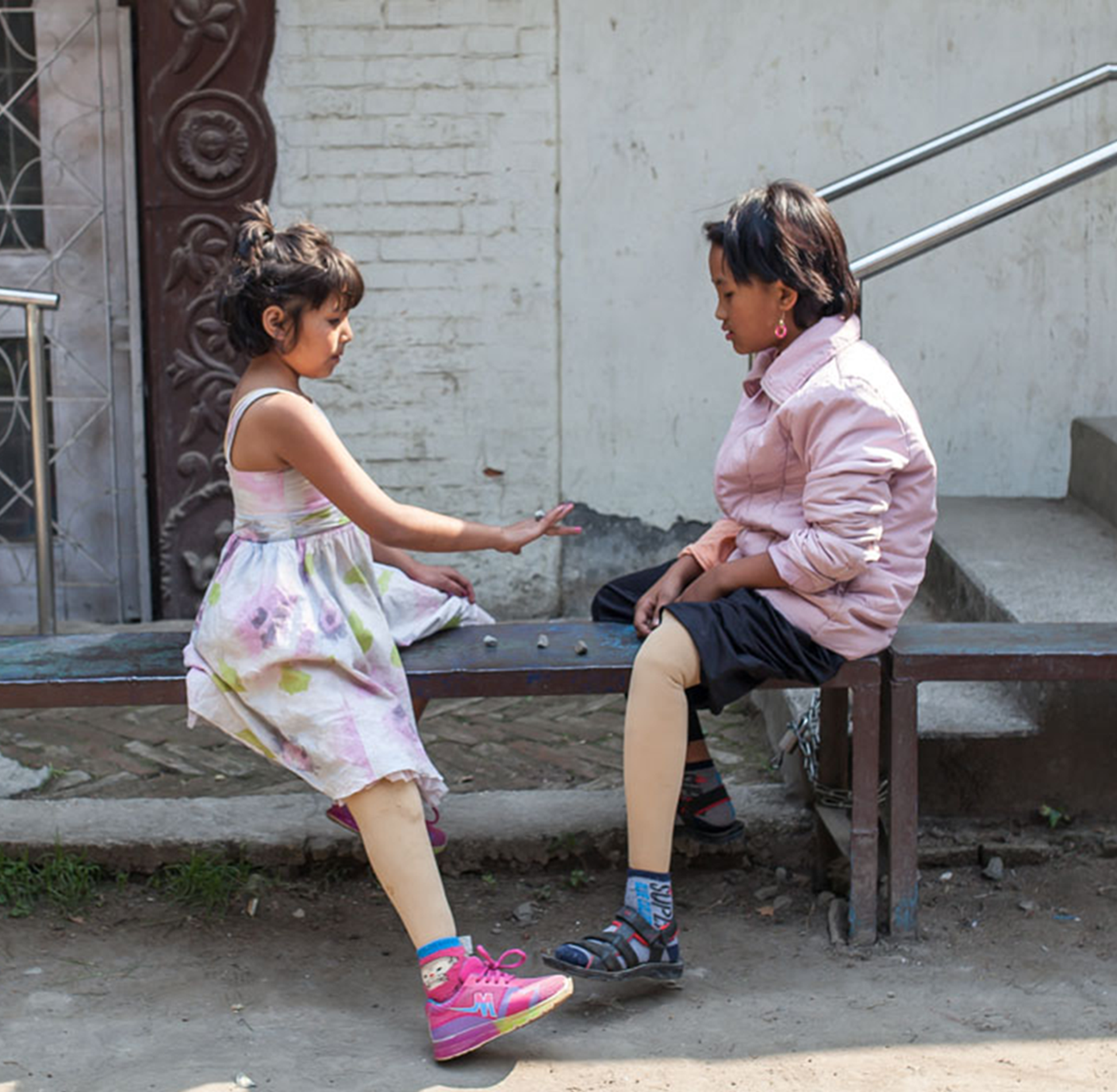 Two children sitting on a bench outdoors, both wearing prosthetic legs, talking together.