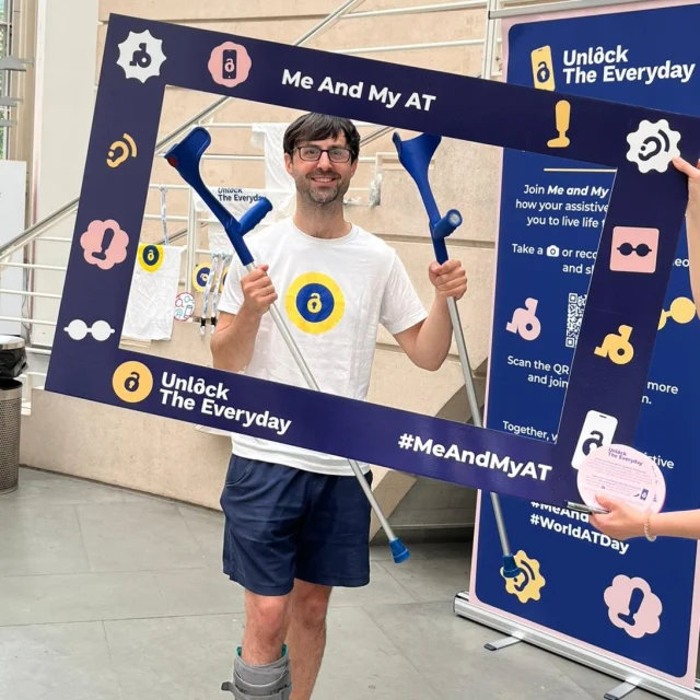 A man stands indoors holding a ‘Me and My AT’ campaign frame and a pair of crutches, with ‘Unlock the Everyday’ and ‘#MeAndMyAT’ text visible.
