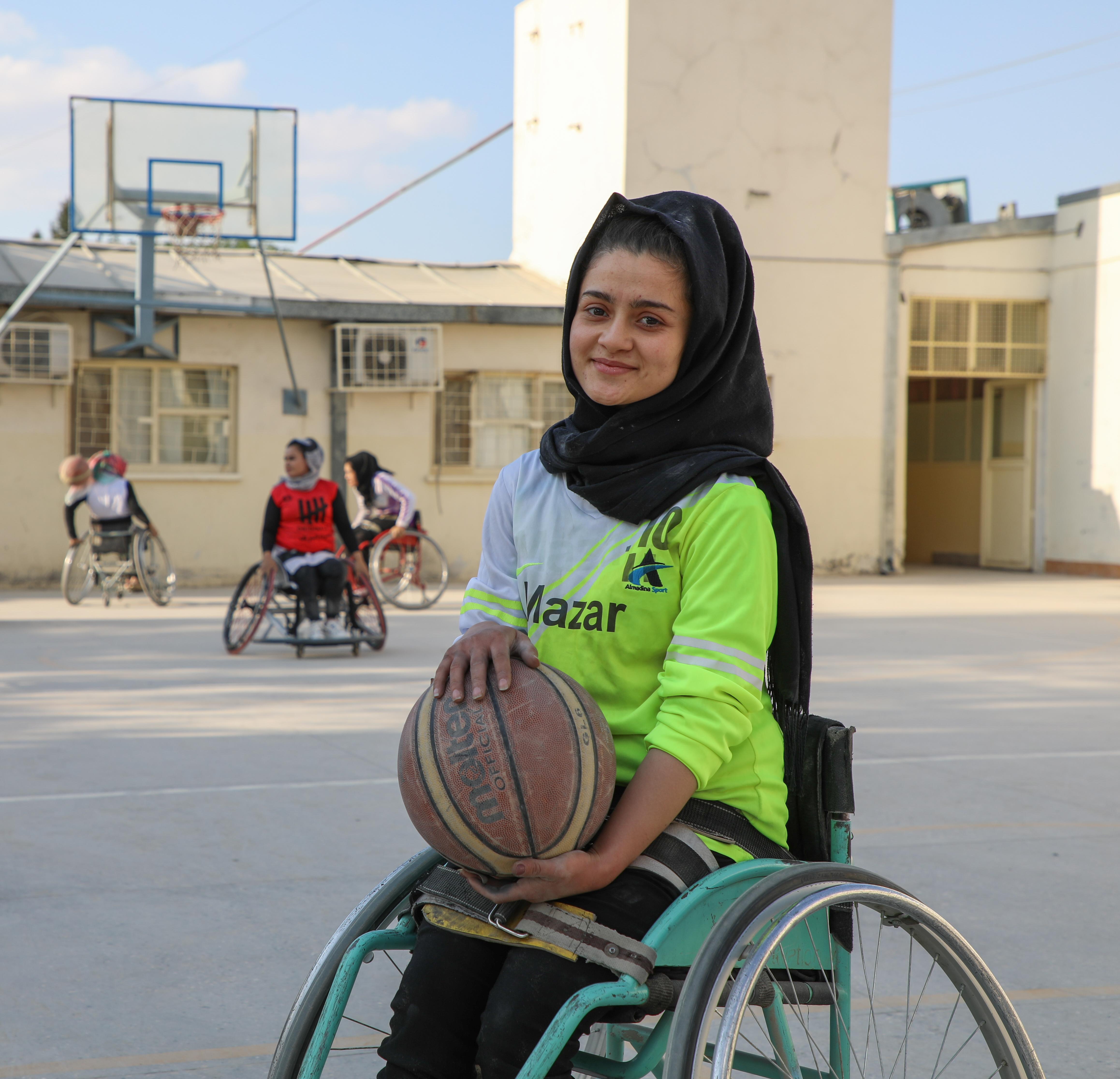 A young woman using a wheelchair holds a basketball on an outdoor court, with others playing wheelchair basketball in the background