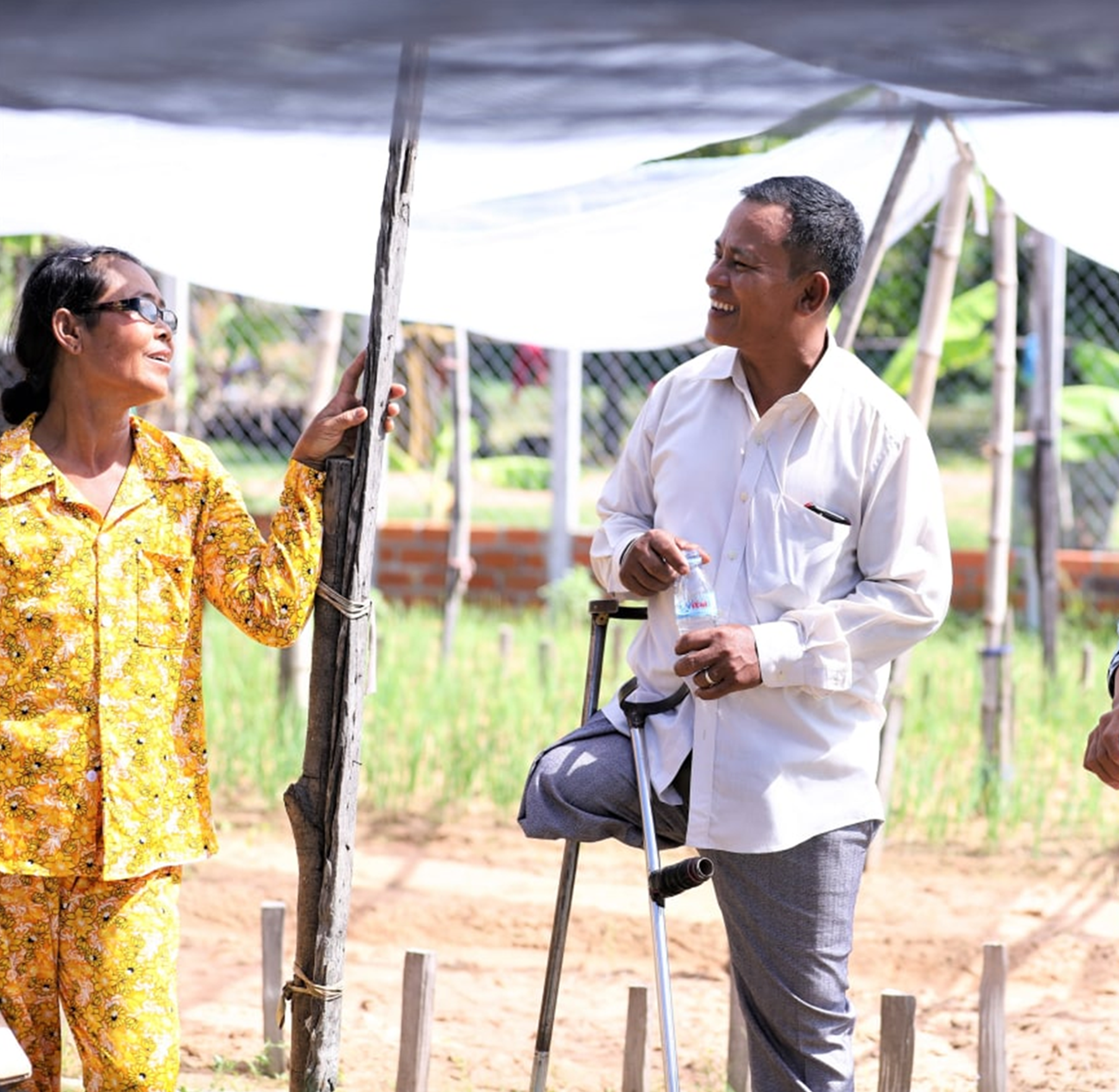 A group of adults talk outdoors under a shaded structure, including a man using a prosthetic leg and a walking aid.