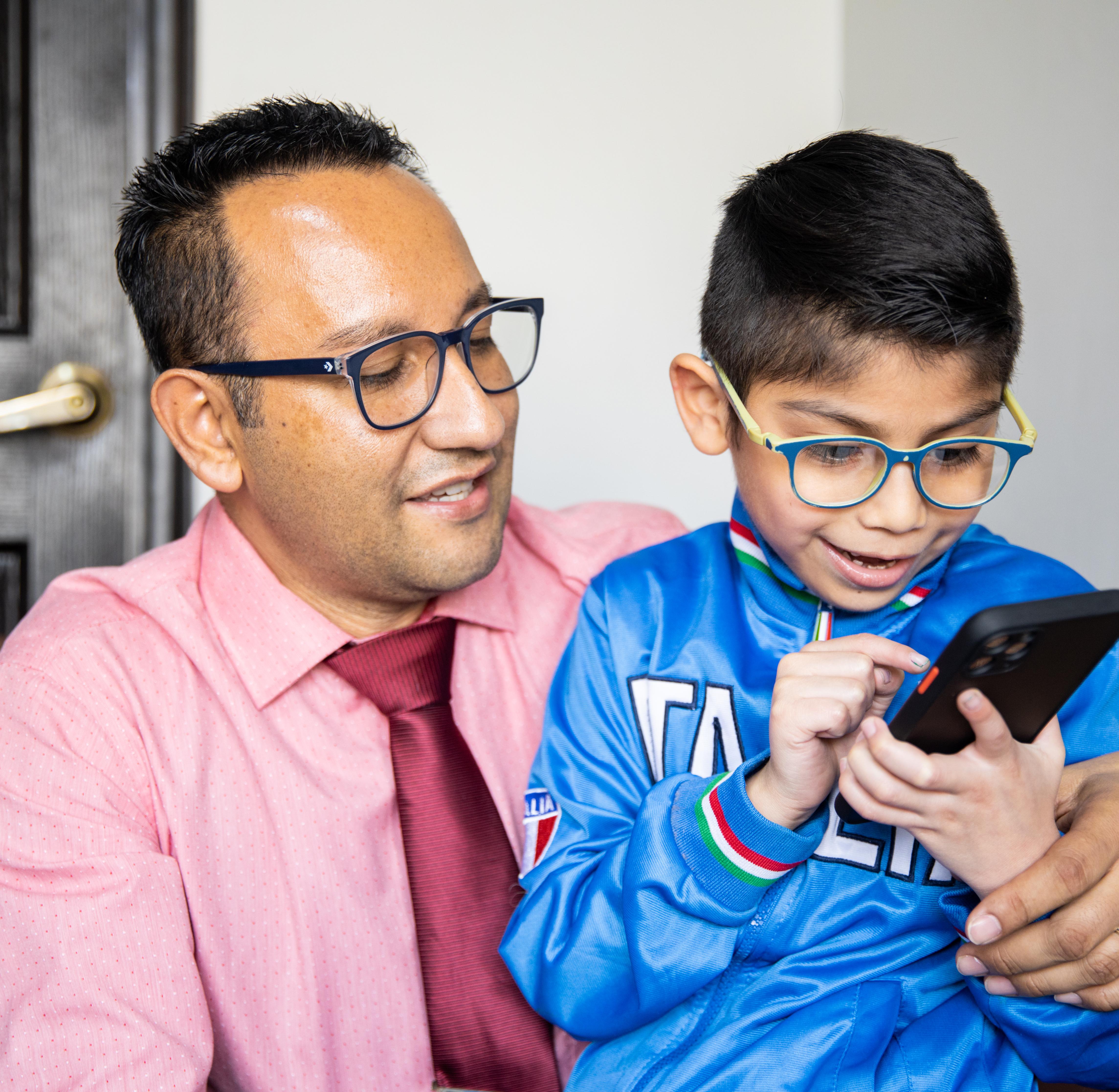 An adult sits with a child who is using a smartphone, both wearing glasses, indoors