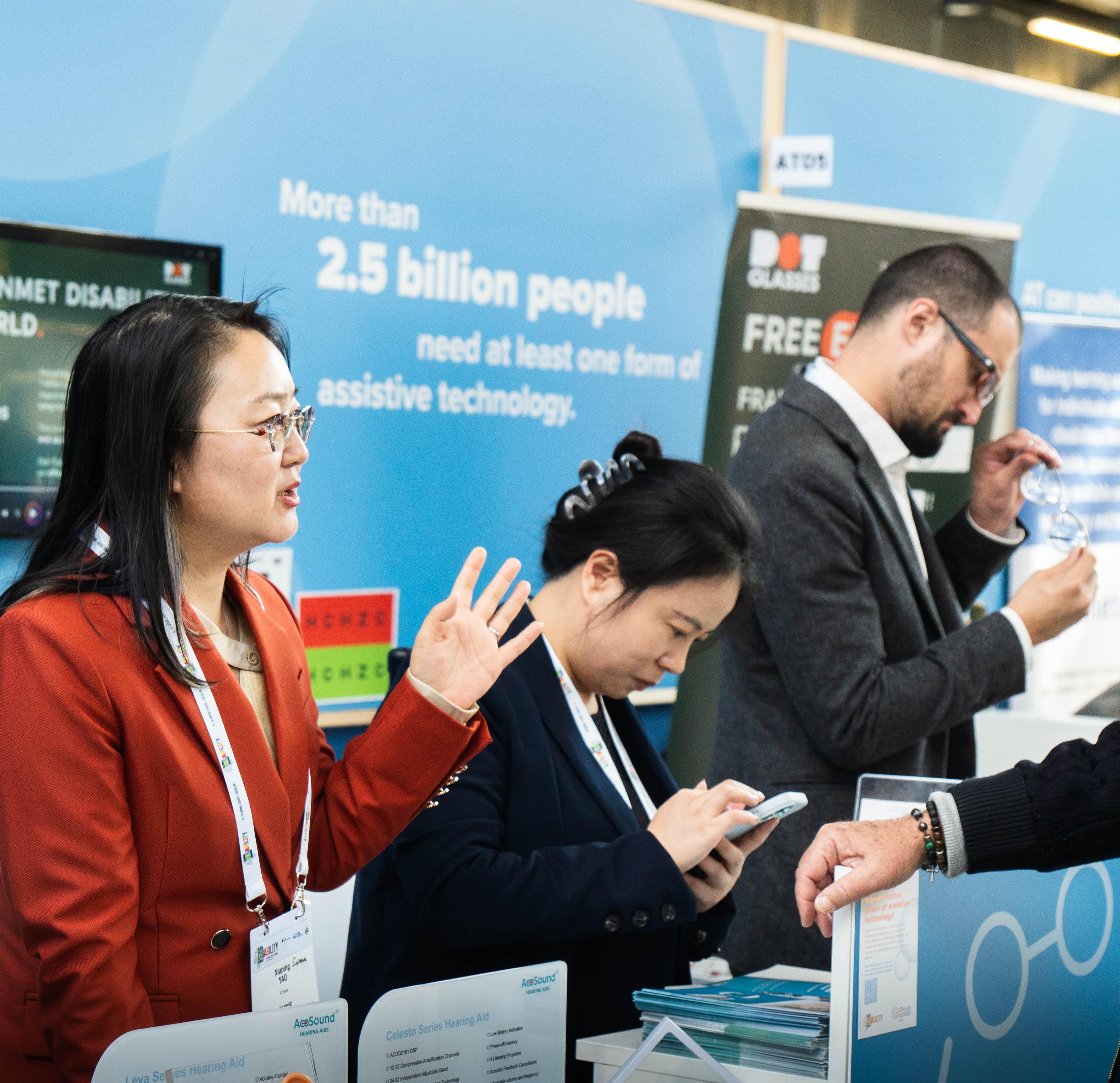 People discuss assistive technology products at an exhibition booth, with informational displays in the background