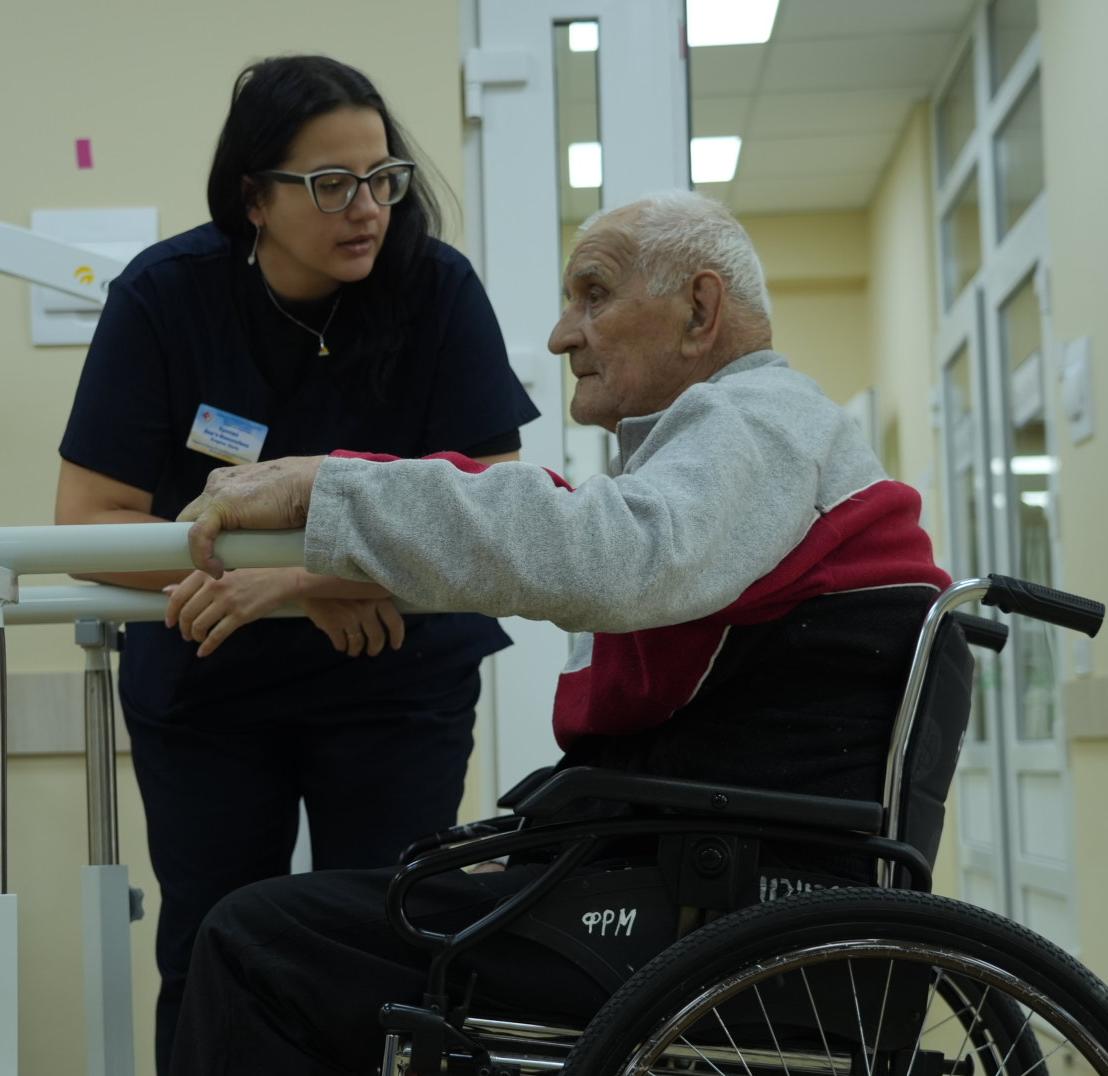 A health professional supports an older man using a wheelchair as he practices standing at parallel bars in a clinic