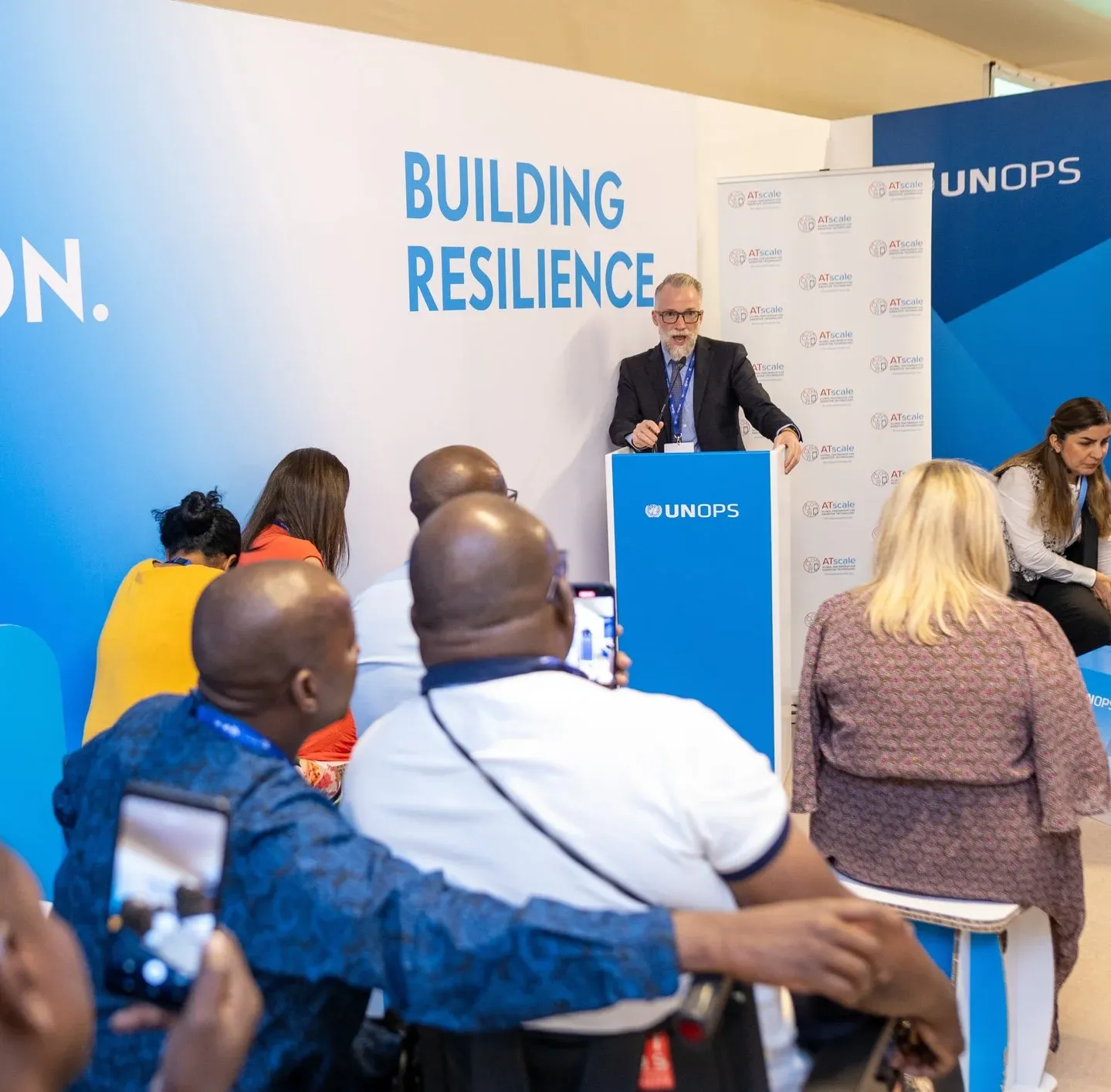 A speaker stands at a UNOPS podium addressing an audience, some using wheelchairs, during a conference session.
