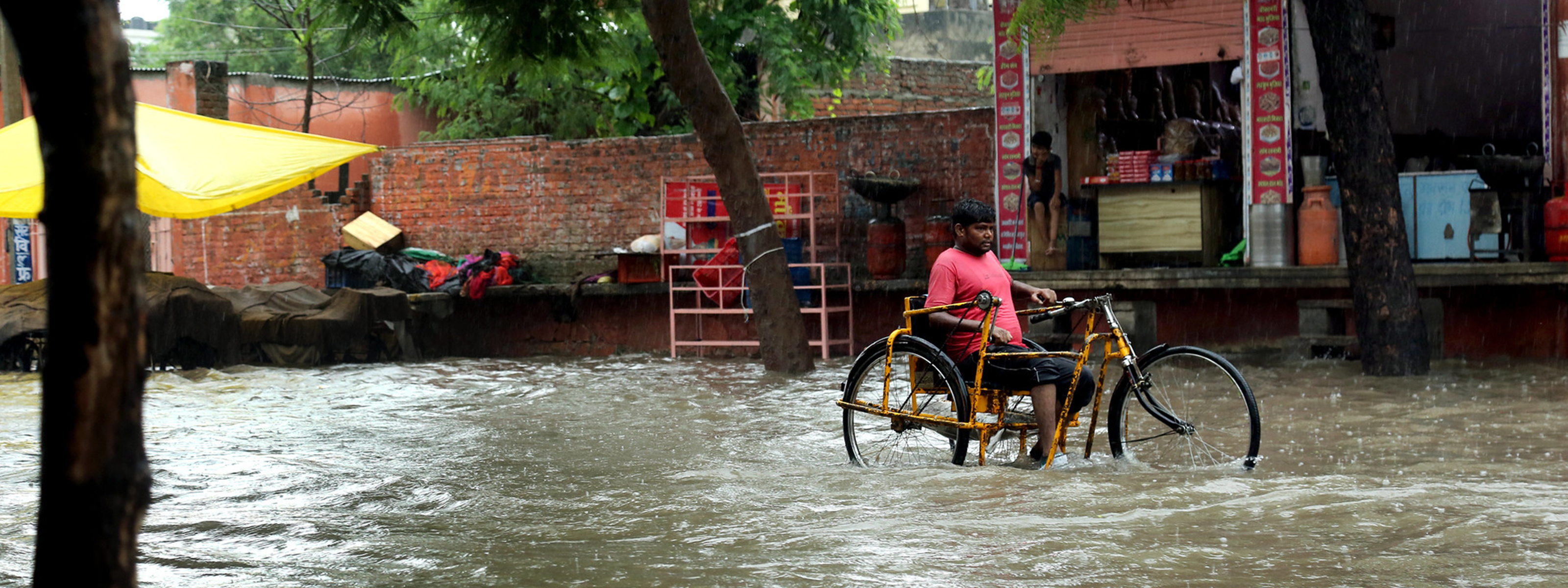 Man using a hand-powered tricycle moves through floodwater on a city street.