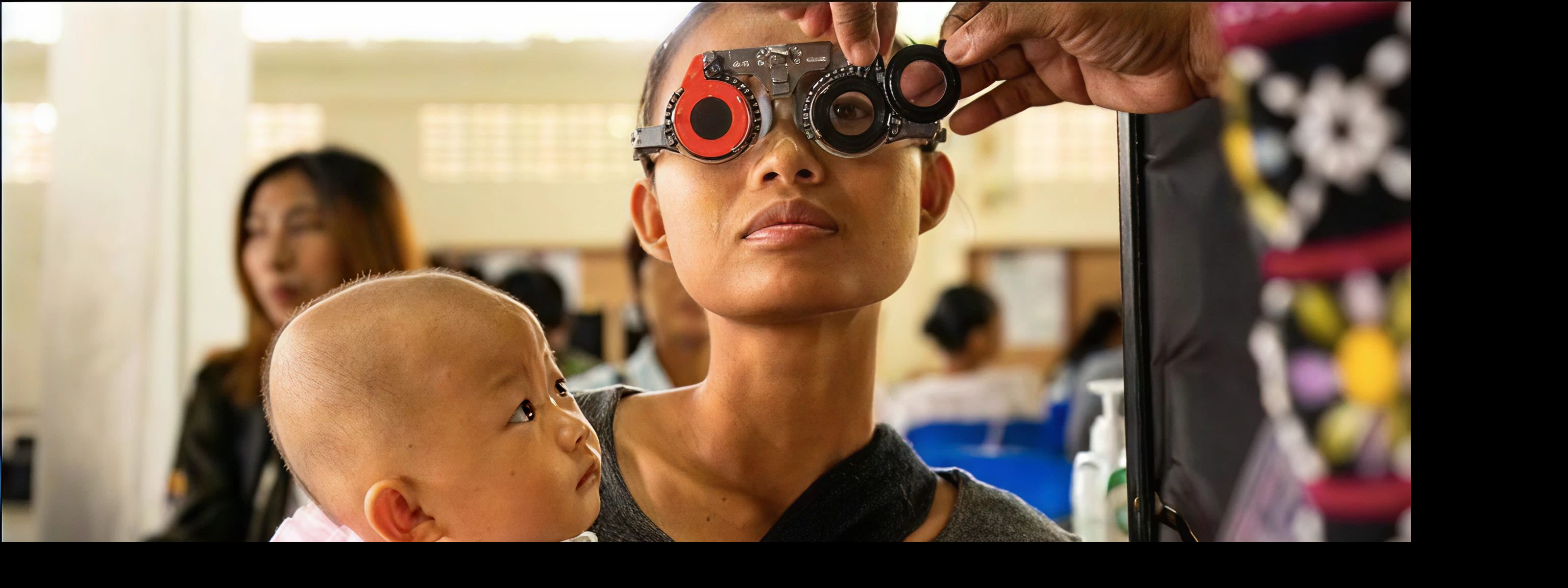 A woman undergoes an eye examination using trial lenses while holding a baby in a clinic setting
