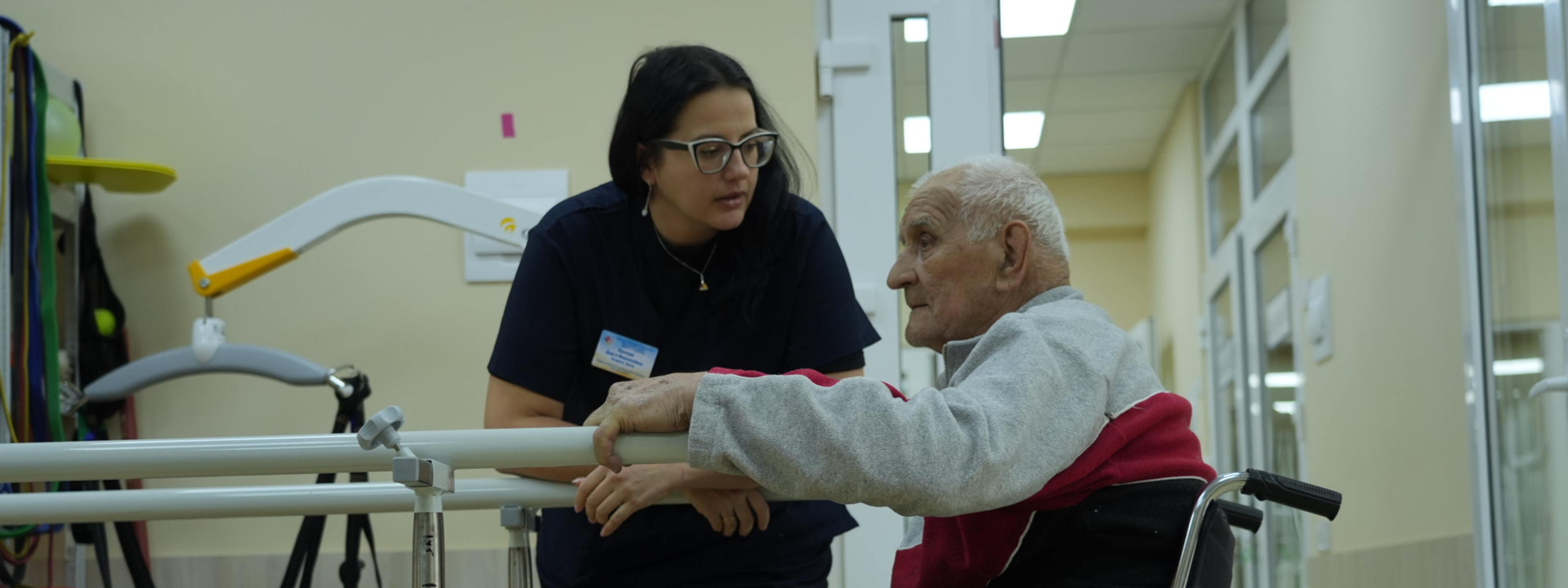 A health professional supports an older man using a wheelchair as he practices standing at parallel bars in a clinic