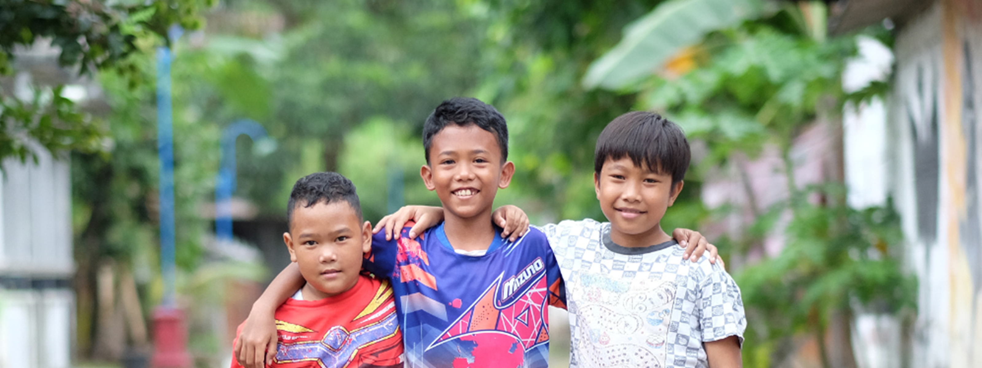 Three boys stand arm in arm on a village path, smiling; the boy in the center uses a lower-limb prosthetic
