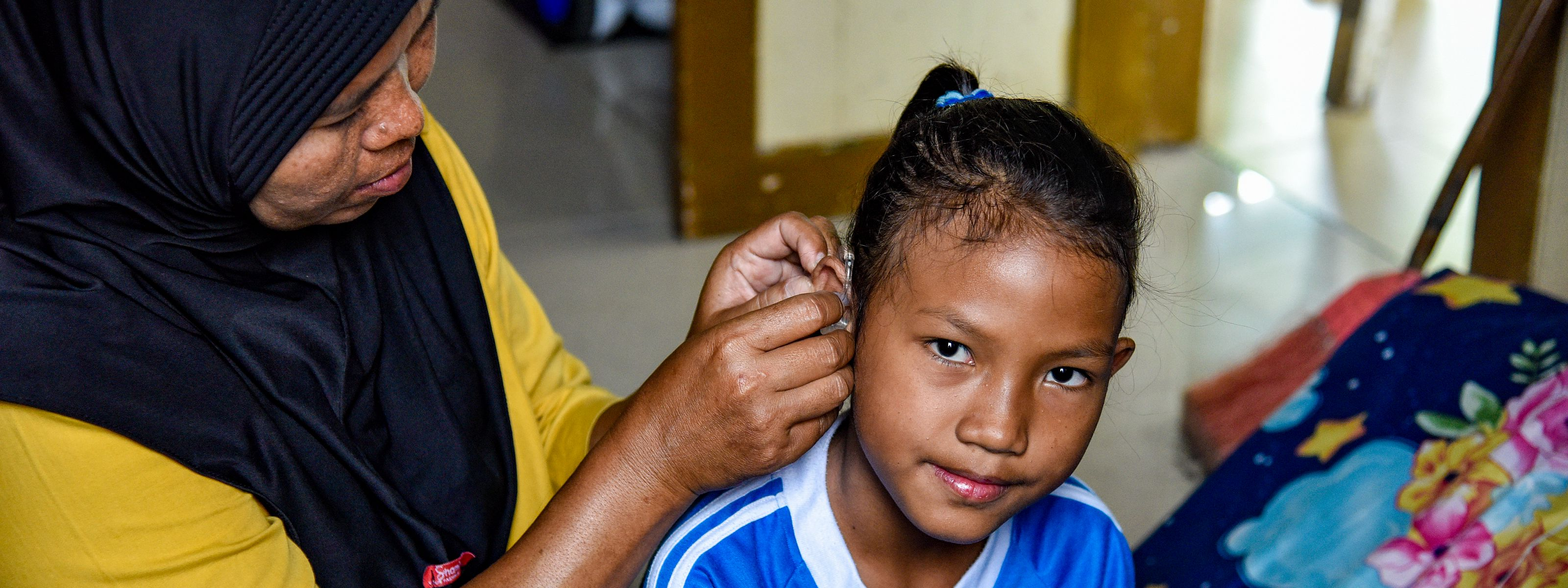 A woman helps a young girl put on a hearing aid indoors