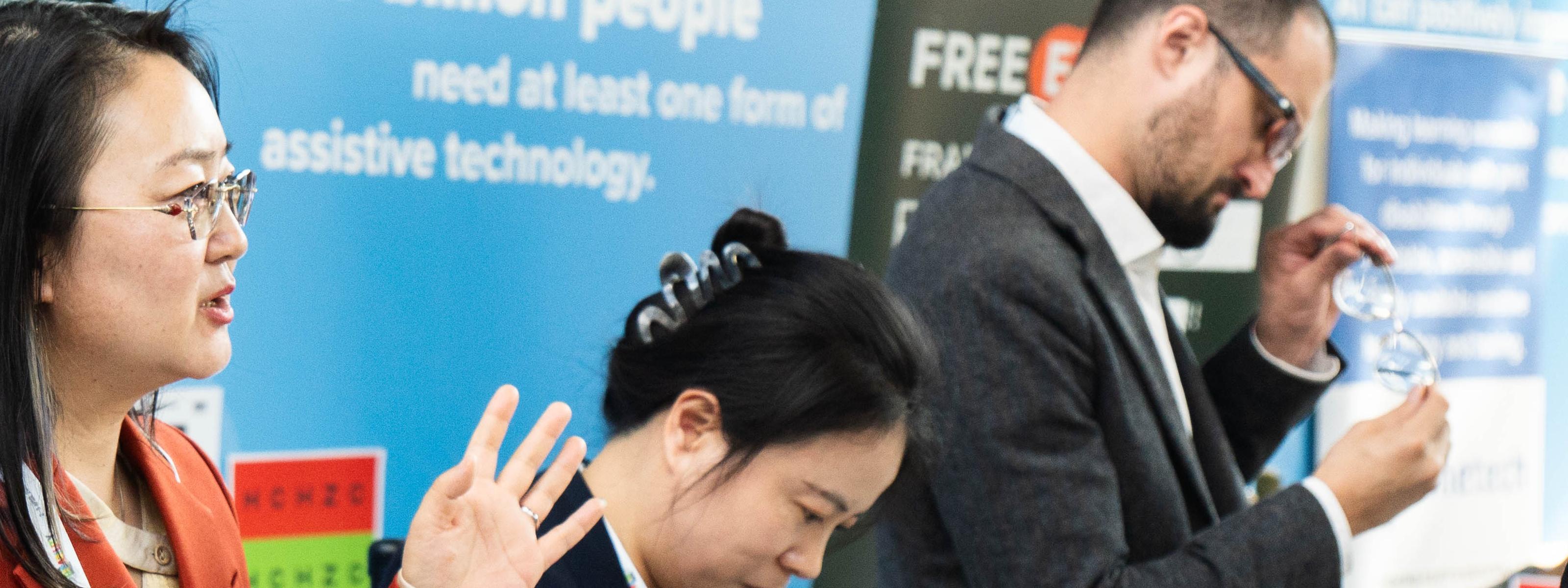 People discuss assistive technology products at an exhibition booth, with informational displays in the background