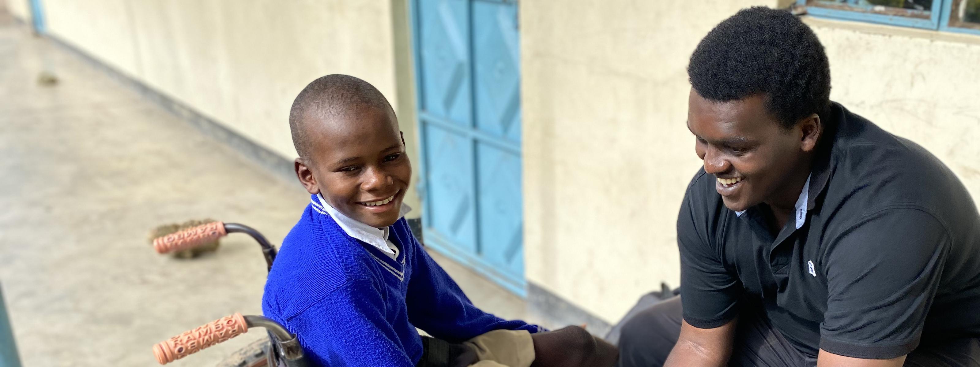 A technician adjusts a wheelchair for a smiling child seated outside a school building.
