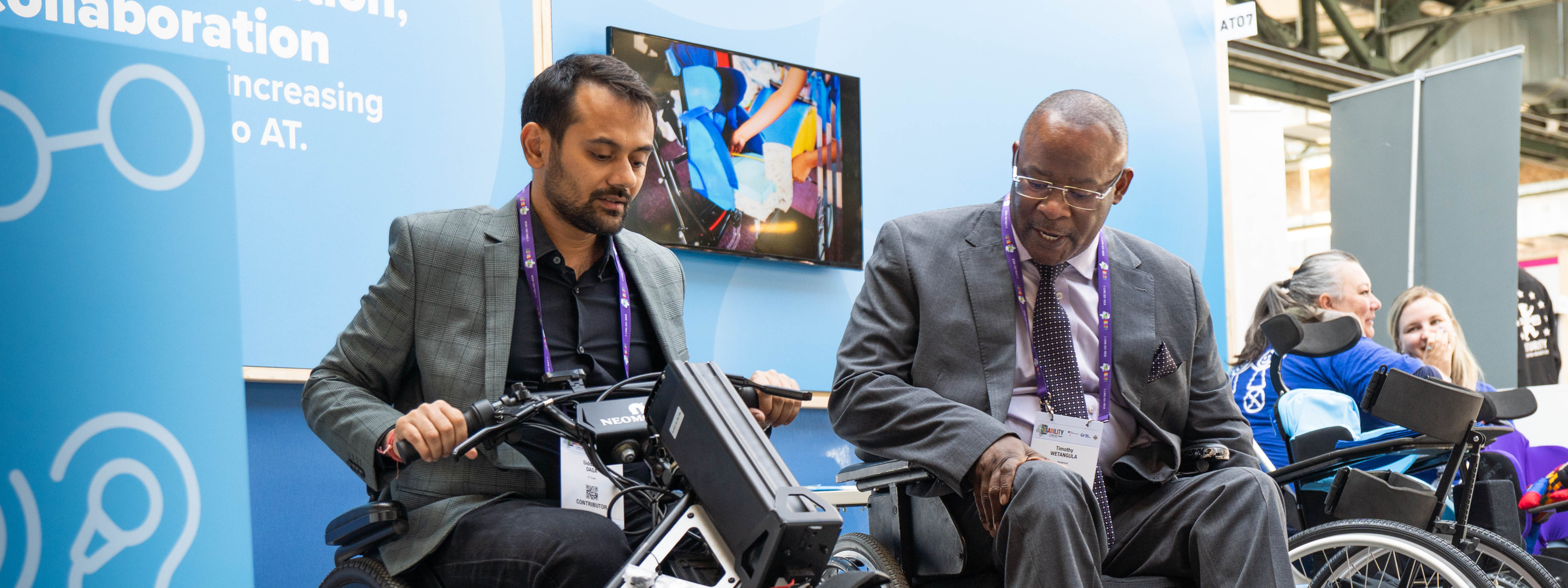 Two men using wheelchairs examine a powered mobility device at an exhibition booth, with other attendees in the background.