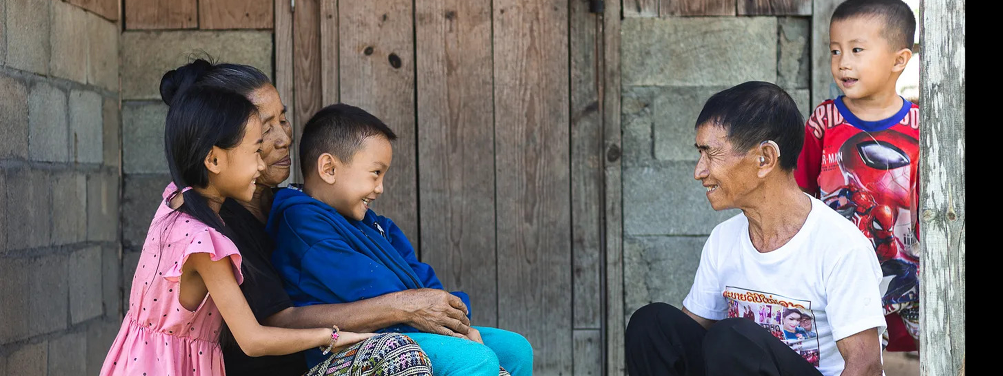 An adult sits outdoors talking with three children seated near a wooden wall.